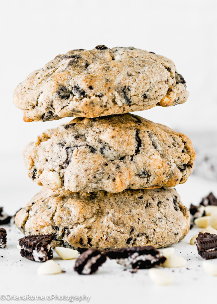 stack of three NYC Bakery-Style Eggless Cookies and Cream Cookies with white chips and chopped Oreos around it over a white surface.