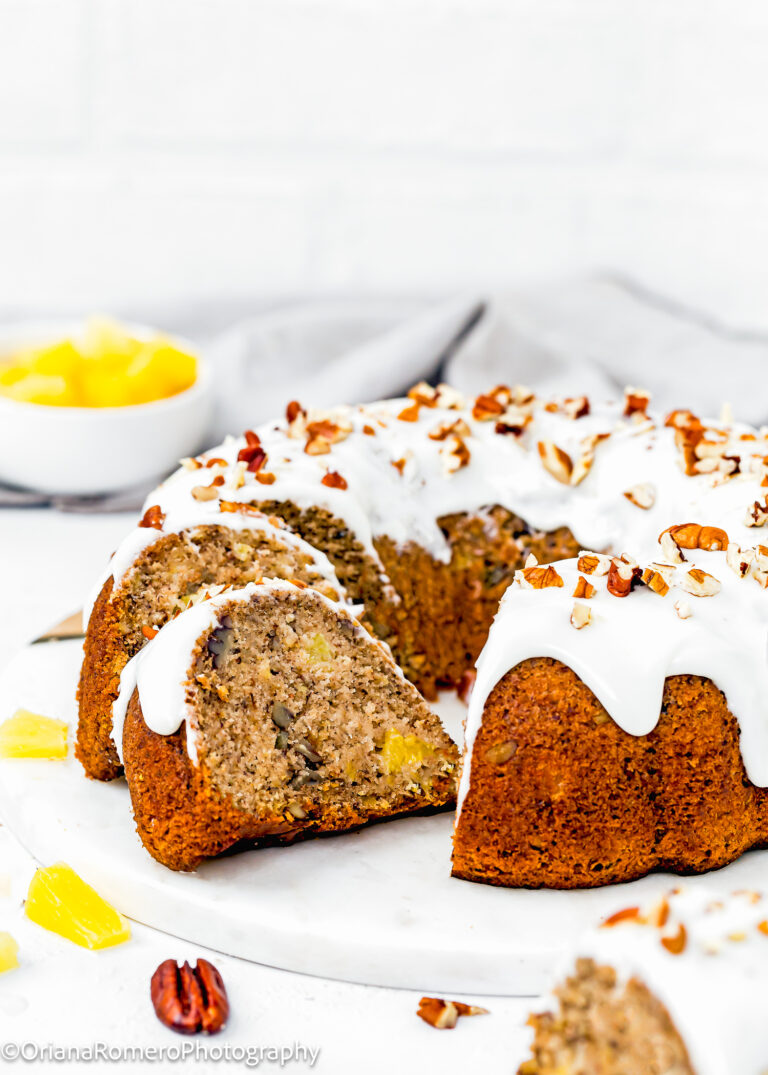 sliced Eggless Hummingbird Bundt Cake with glaze and chopped pecans on top on a serving plate with more pecan and pineapple pieces on the side.